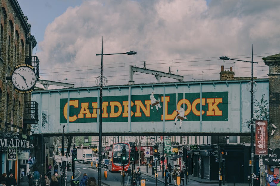 A large, weathered metal bridge with the words 'Camden Lock' painted in bold yellow and red letters spans above a busy street in Camden Market. The bridge is supported by metal beams and has electrica