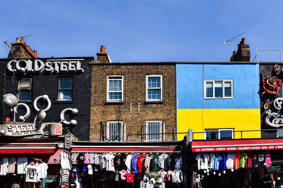A large, weathered metal bridge with the words 'Camden Lock' painted in bold yellow and red letters spans above a busy street in Camden Market. The bridge is supported by metal beams and has electrica