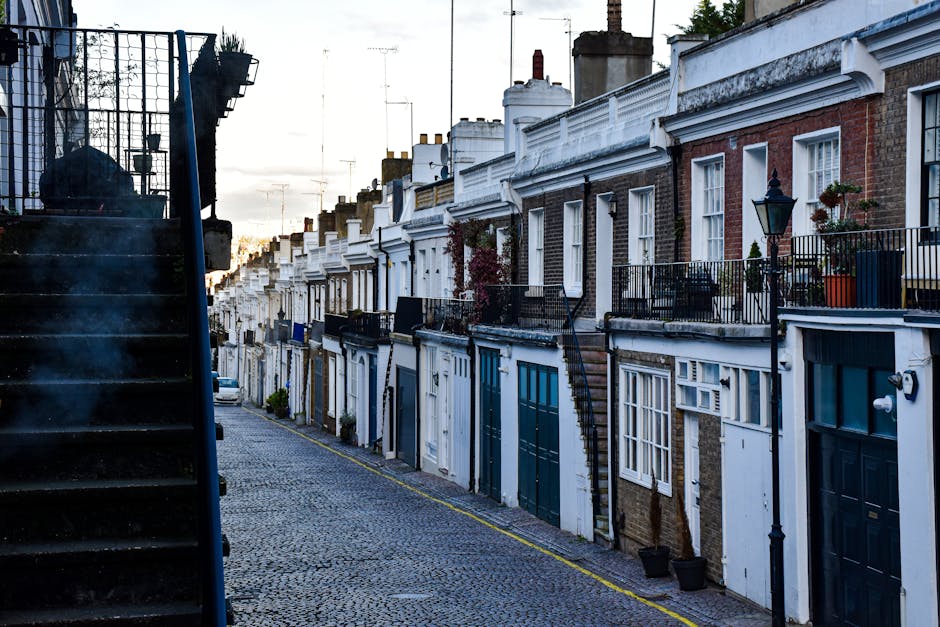 A narrow residential street in Mornington Crescent with terraced brick houses featuring white decorative facades, black metal railings, and small front gardens with potted plants. On the left side, th